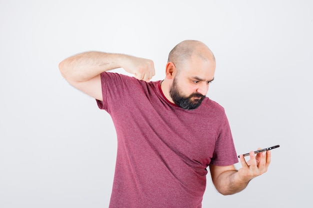 Junger Mann, der auf das Telefon schaut und die Faust im rosa T-Shirt ballt und wütend aussieht. Vorderansicht.