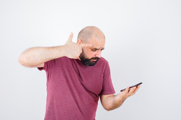 Junger mann, der auf das telefon schaut und eine waffengeste in rosa t-shirt zeigt und düster aussieht. vorderansicht.