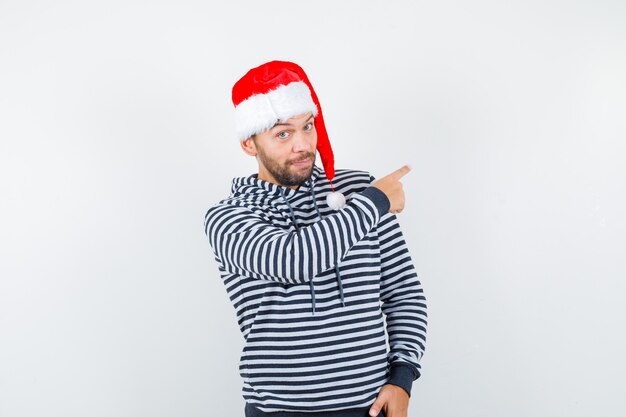 Junger Mann, der auf die obere rechte Ecke in Hoodie, Weihnachtsmütze zeigt und selbstbewusst aussieht,