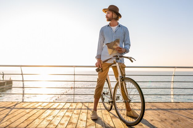 Junger Mann, der auf Fahrrad durch See in den Sommerferien durch das Meer auf Sonnenuntergang reist und Kartensicht mit Kamera hält