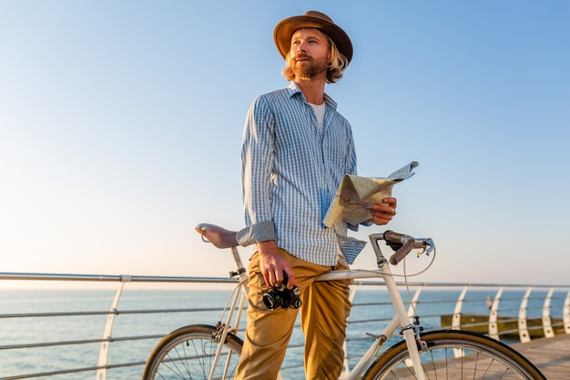 Junger Mann, der auf Fahrrad durch See in den Sommerferien durch das Meer auf Sonnenuntergang reist und Kartensicht mit Kamera hält