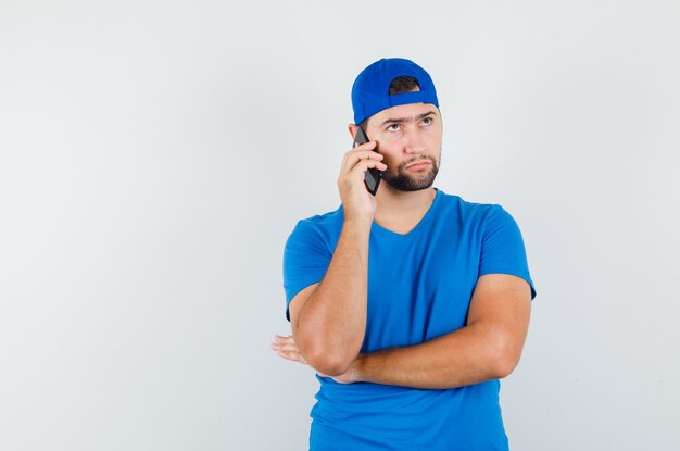 Junger Mann, der auf Handy in blauem T-Shirt und Mütze spricht und ernst schaut