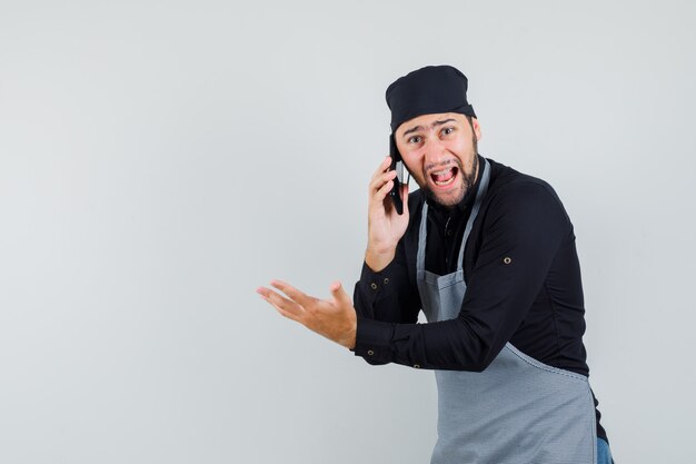 Junger Mann, der auf Handy in Hemd, Schürze spricht und wütend schaut, Vorderansicht.