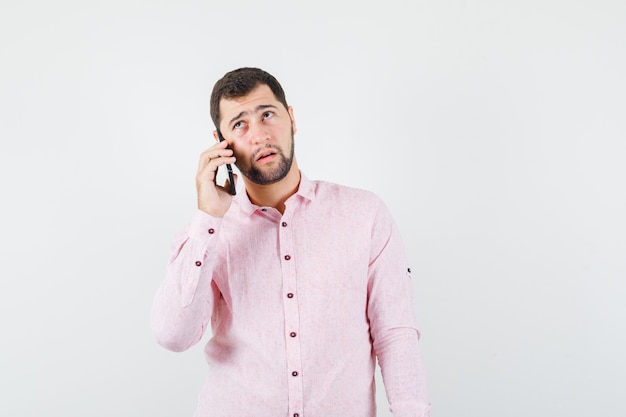 Junger Mann, der auf Handy in rosa Hemd spricht und nachdenklich schaut