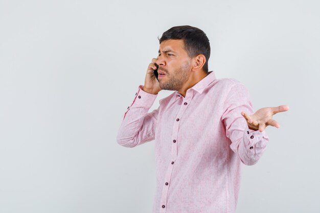 Junger Mann, der auf Handy in rosa Hemd spricht und nervöse Vorderansicht schaut.