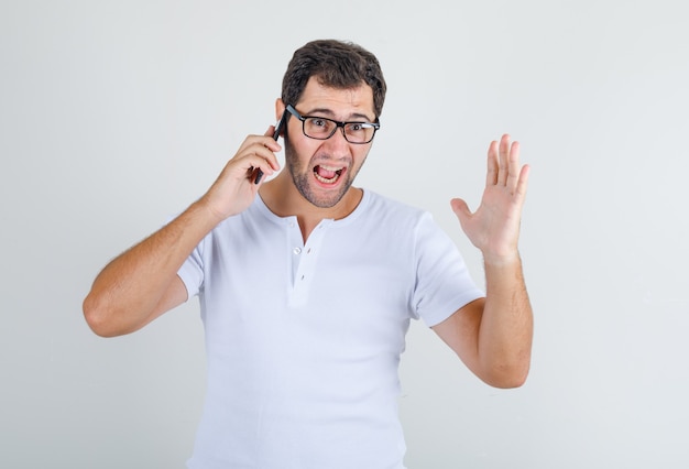 Junger Mann, der auf Handy in weißem T-Shirt, Brille schreit und gereizt schaut