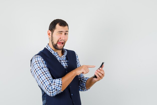 Junger Mann, der auf Handy zeigt, Augenzwinkern, Zunge in Hemd, Weste, Vorderansicht herausstrecken.