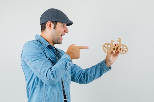 Junger mann, der auf hölzernes spielzeugfahrrad in der kappe, im t-shirt, in der jacke zeigt und glücklich schaut. .