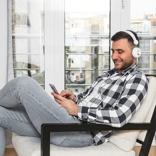 Junger Mann, der auf hörender Musik des Stuhls am Kopfhörer durch Handy sitzt