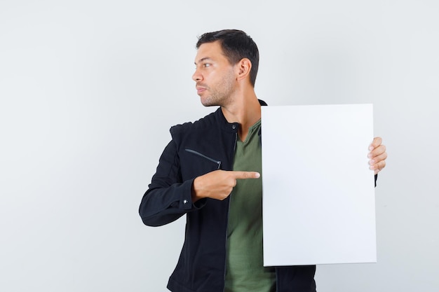 Junger Mann, der auf leere Leinwand zeigt, während er in T-Shirt, Jacke und konzentriertem Blick beiseite schaut. Vorderansicht.