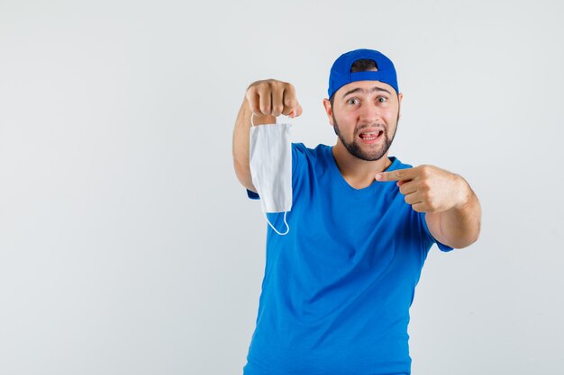 Junger Mann, der auf medizinische Maske im blauen T-Shirt und in der Kappe zeigt und vorsichtig schaut