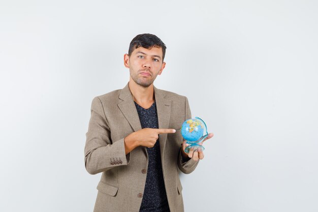 Junger Mann, der auf Mini-Globus in graubrauner Jacke, schwarzem Hemd zeigt und fokussiert aussieht, Vorderansicht.