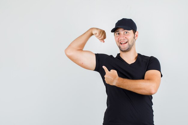 Junger Mann, der auf Muskel in schwarzem T-Shirt, Mütze zeigt und fröhlich schaut