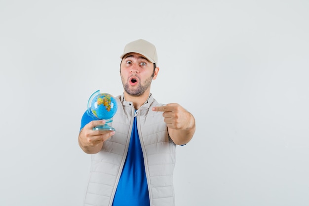 Junger Mann, der auf Schulkugel im T-Shirt, in der Jacke zeigt und erstaunt schaut. Vorderansicht.