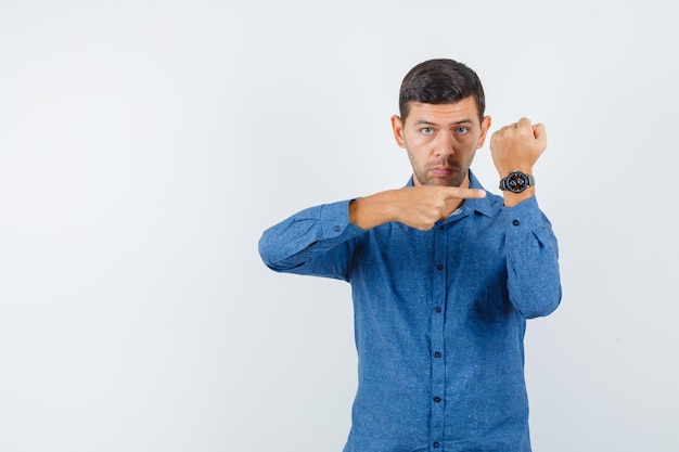 Junger Mann, der auf seine Uhr im blauen Hemd zeigt und pünktlich aussieht. Vorderansicht.