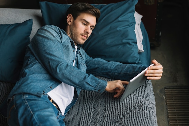 Junger Mann, der auf Seite über dem Bett unter Verwendung der digitalen Tablette liegt