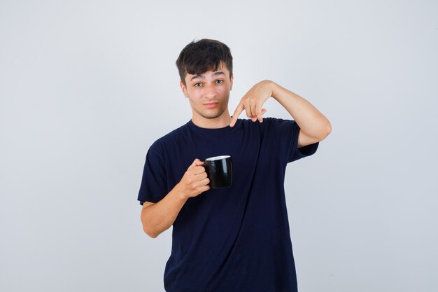 Junger Mann, der auf Tasse Tee im schwarzen T-Shirt zeigt und verwirrt schaut. Vorderansicht.