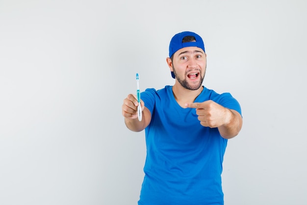 Junger Mann, der auf Zahnbürste im blauen T-Shirt und in der Kappe zeigt und positiv schaut