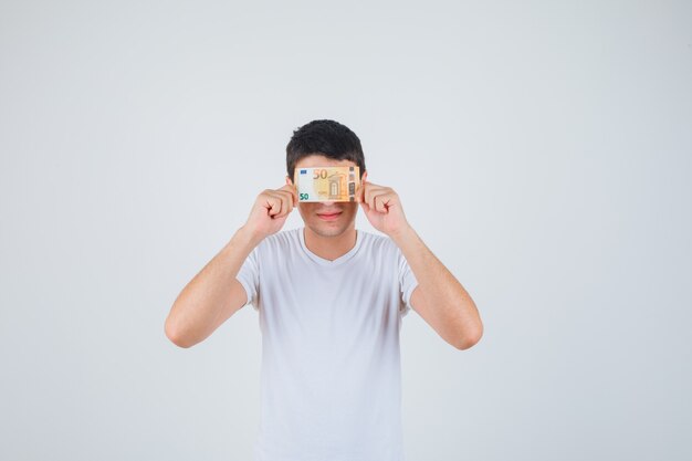 Junger Mann, der Auge mit Euro-Banknote im T-Shirt bedeckt und fröhlich schaut. Vorderansicht.