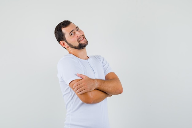 Junger Mann, der beim Stehen verschränkte Arme im T-Shirt aufwirft und fröhlich schaut. Vorderansicht.