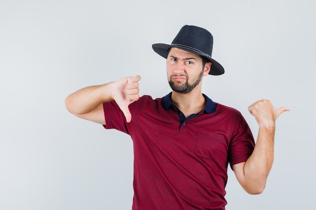 Junger Mann, der beiseite zeigt, während Daumen unten in T-Shirt, Hut zeigt und unzufrieden schaut, Vorderansicht.