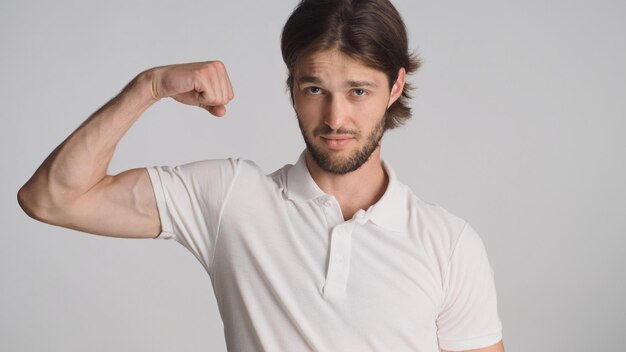 Junger Mann, der Bizeps zeigt, der auf weißem Hintergrund zuversichtlich aussieht Sportlicher Kerl, der Körperstärke in die Kamera demonstriert