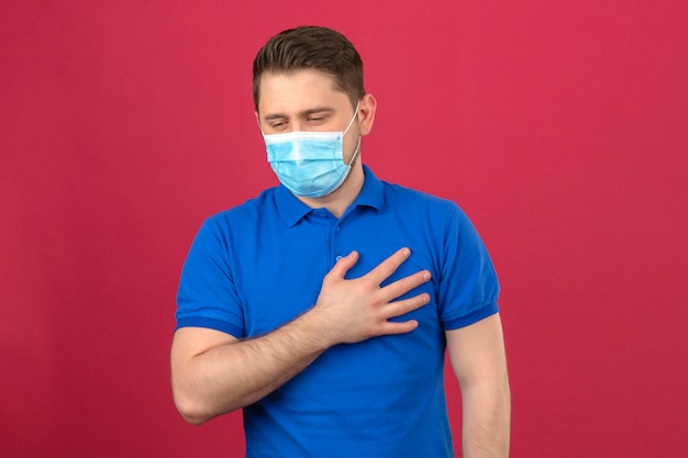 Junger Mann, der blaues Poloshirt in der medizinischen Schutzmaske trägt, die seine Hand auf Brustlungen hält, die sich schlecht fühlen, über isolierter rosa Wand zu stehen