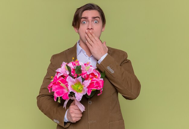 Junger Mann, der Blumenstrauß hält, der Kamera betrachtet, die schockiert ist, Mund bedeckt