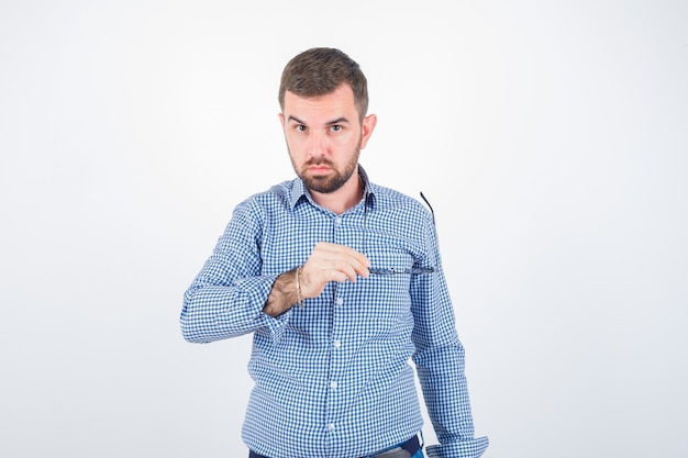 Junger Mann, der Brille hält, während Kamera in Hemd, Jeans und gutaussehend schaut. Vorderansicht.