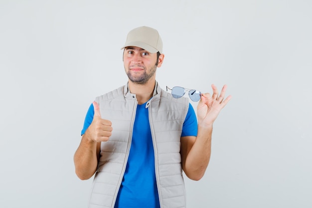 Junger Mann, der Brille hält, zeigt Daumen oben in T-Shirt, Jacke, Mütze, Vorderansicht.