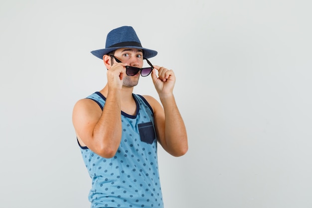 Junger Mann, der Brille im blauen Unterhemd, im Hut abnimmt und konzentriert schaut. Vorderansicht.