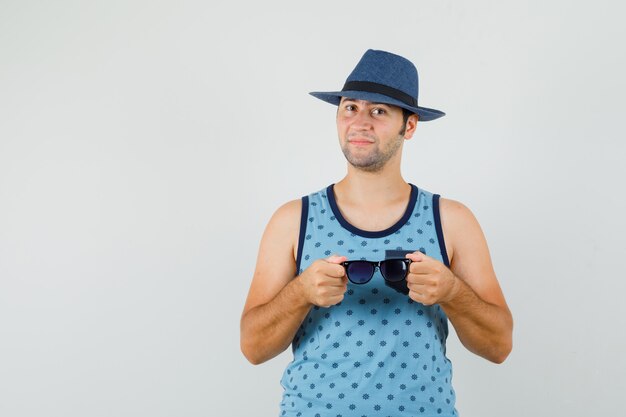 Junger Mann, der Brille im blauen Unterhemd, im Hut hält und zuversichtlich schaut.