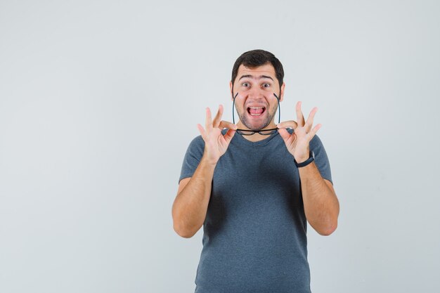 Junger Mann, der Brille im grauen T-Shirt abnimmt und glücklich schaut