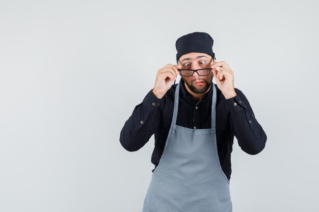 Junger Mann, der Brille mit zusammengekniffenen Augen im Hemd, in der Schürze abnimmt und lustig schaut. Vorderansicht.