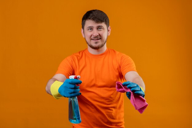 Junger Mann, der das orangefarbene T-Shirt hält, das Reinigungsspray und Teppich hält, der fröhlich positiv und glücklich betrachtet Kamera genießt, bereit zu stehen, über orange Hintergrund zu stehen