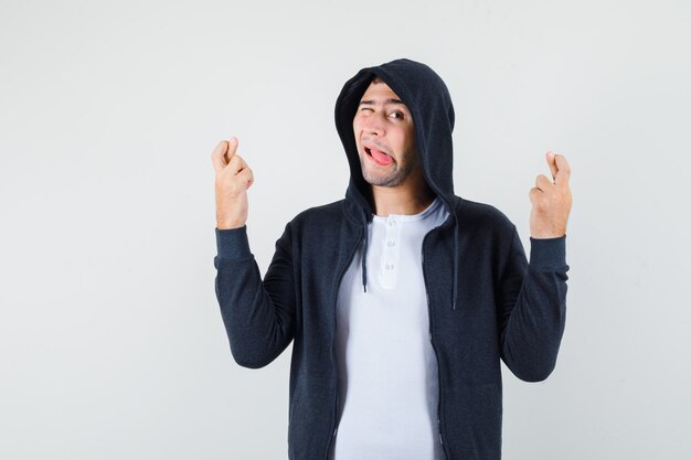 Junger Mann, der Daumen drückt, Auge zwinkert, Zunge in T-Shirt, Jacke, Vorderansicht herausstreckt.