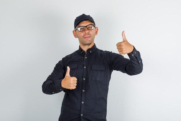 Junger Mann, der Daumen oben im schwarzen Hemd mit Kappe, Brille zeigt