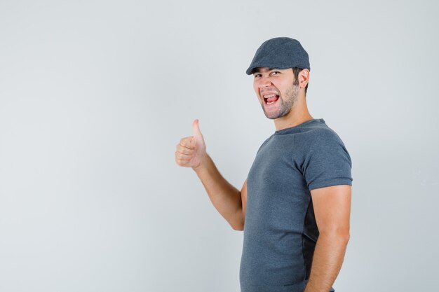 Junger Mann, der Daumen oben in grauer T-Shirt-Kappe zeigt und glücklich schaut