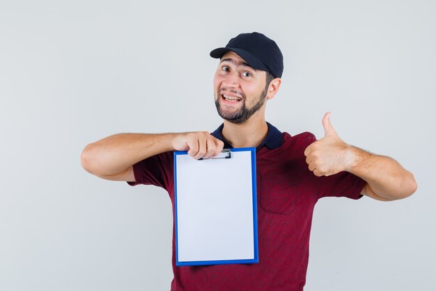 Junger Mann, der Daumen oben zeigt, während Notizbuch im roten T-Shirt, schwarze Kappe zeigt und glücklich schaut. Vorderansicht.