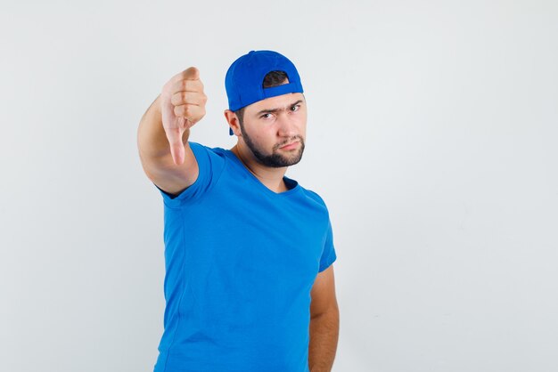 Junger Mann, der Daumen unten im blauen T-Shirt und in der Kappe zeigt und düster aussieht