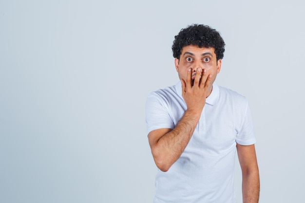 Junger Mann, der den Mund mit der Hand im weißen T-Shirt und in den Jeans bedeckt und überrascht schaut. Vorderansicht.