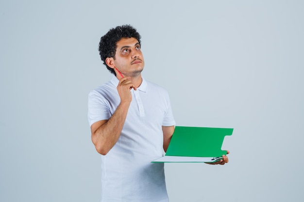 Junger Mann, der den Stift in der Heureka-Geste anhebt und ein Notizbuch in weißem T-Shirt und Jeans hält und nachdenklich aussieht. Vorderansicht.