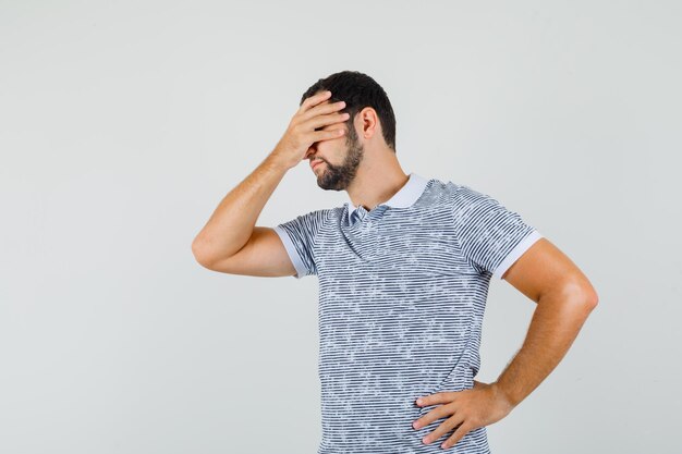 Junger Mann, der die Hand auf den Augen im T-Shirt hält und verlegen schaut. Vorderansicht.