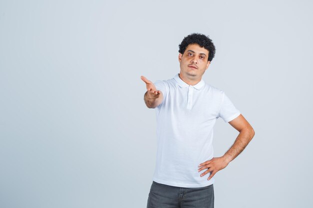 Junger Mann, der die Hand in verwirrter Geste in weißem T-Shirt, Hose streckt und selbstbewusst aussieht. Vorderansicht.