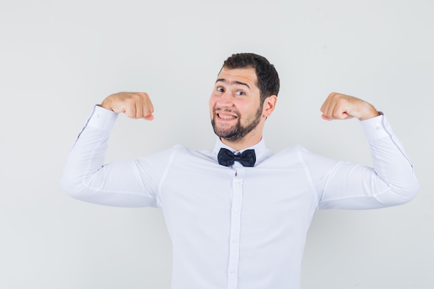 Junger Mann, der die Muskeln der Arme im weißen Hemd zeigt und glücklich schaut. Vorderansicht.