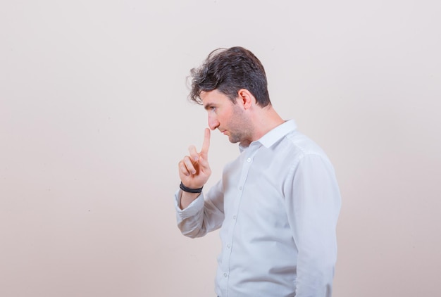 Junger Mann, der die Nase mit dem Finger im Hemd berührt und nachdenklich aussieht