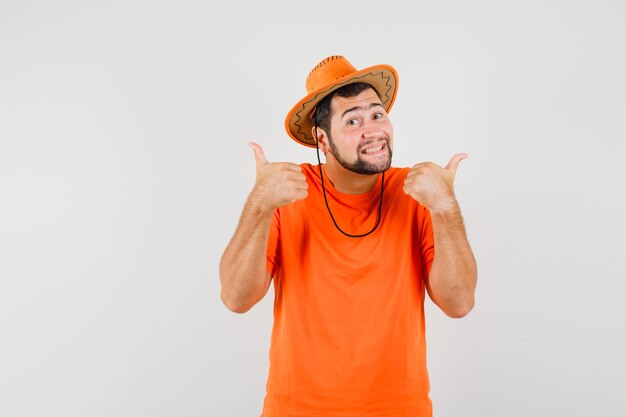 Junger Mann, der doppelte Daumen oben in orange T-Shirt, Hut und lustig aussehend, Vorderansicht zeigt.