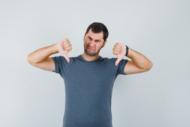 Junger Mann, der doppelte Daumen unten im grauen T-Shirt zeigt und niedergeschlagen aussieht. Vorderansicht.