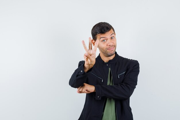 Junger Mann, der drei Finger im T-Shirt, in der Jacke und in der fröhlichen Vorderansicht hält.