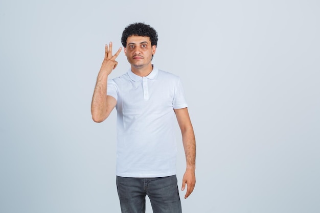 Junger Mann, der drei Finger im weißen T-Shirt, in der Hose zeigt und selbstbewusst aussieht, Vorderansicht.